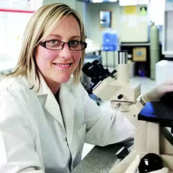 Woman wearing glasses and a white lab coat sitting in front of lab equipment and smile for the camera. New equipment brings cancer's complexity into focus