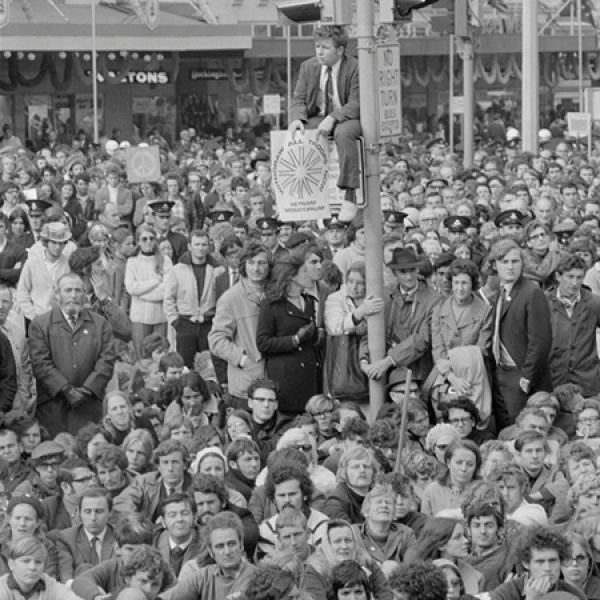 A large crowd fills a city street during a protest, with many people seated on the ground, others standing tightly packed together, and one individual perched high on a pole above the crowd near visible protest signs.