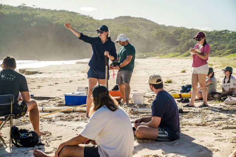 Students sit and stand on the sand, listening to an Academic staff member talk about the beach.