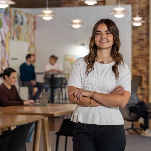 Girl inside building in white shirt, black pants and smiling at camera with arms crossed across chest.
