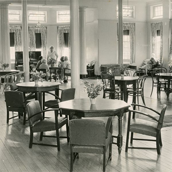Black-and-white photograph of a spacious hall with wooden tables and chairs, where a small group sits around one table while another person reads by a window, with tall columns and high windows letting in soft light.