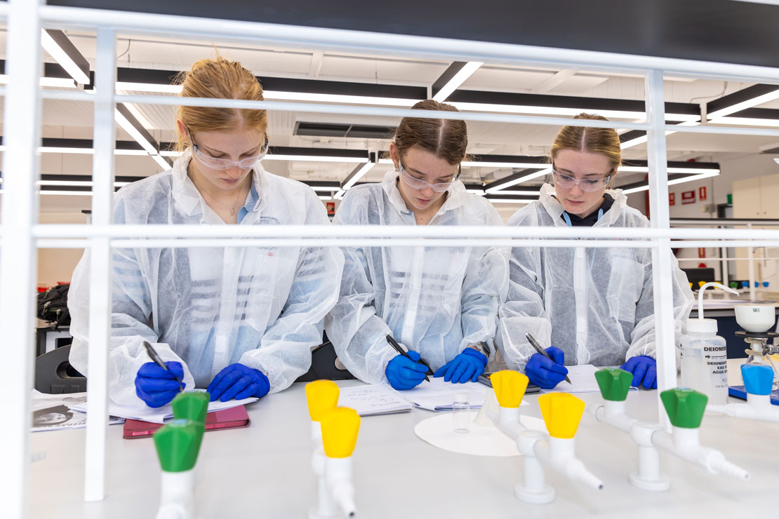 Three high school students stand in a lab, writing notes, wearing lab coats and protective glasses