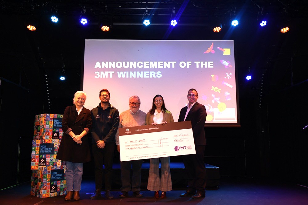 Woman holding a novelty cheque with three men and another woman standing beside her, all smiling for the camera