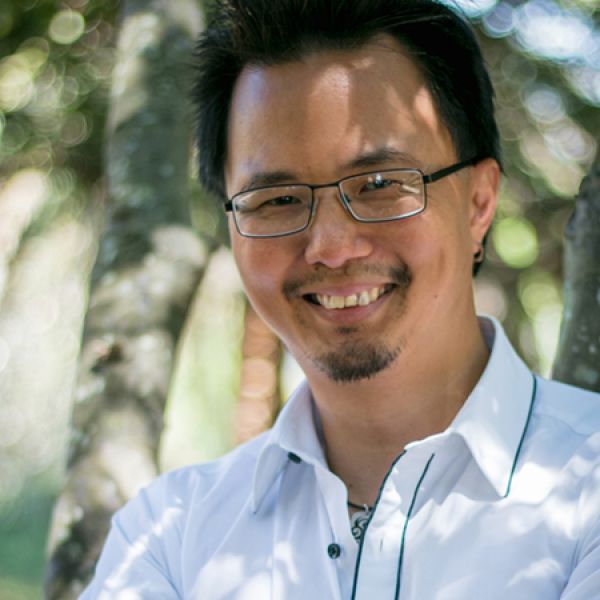 Close-up outdoor portrait of a person wearing rectangular glasses and a white collared shirt, with trees and softly blurred greenery in the background.