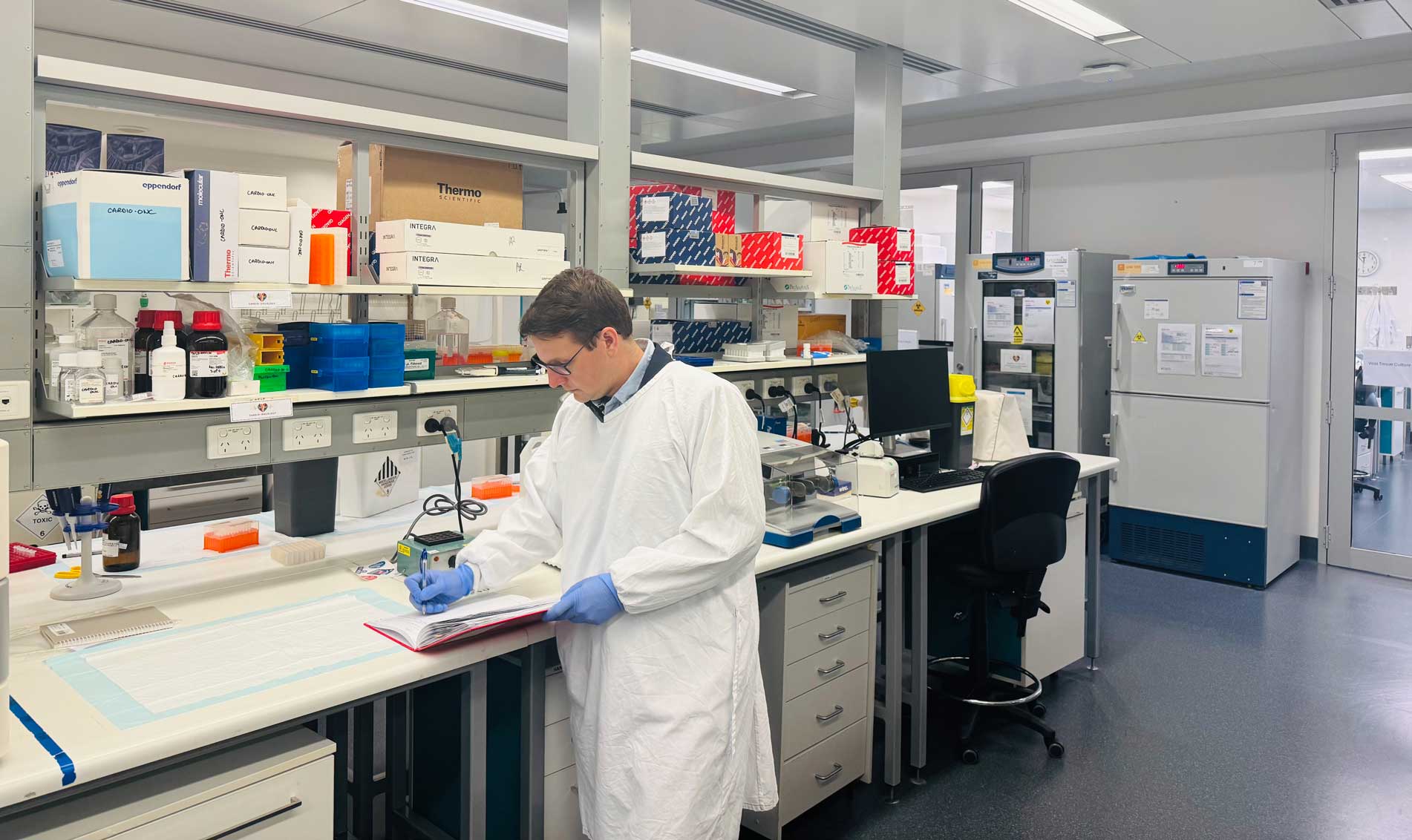 Researcher, Associate Professor Gerard Kaiko, wearing a laboratory gown and gloves records observations in a notebook at a laboratory workstation surrounded by scientific equipment and supplies.