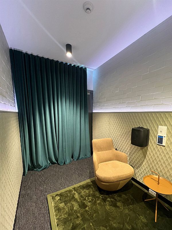 A brown lounge chair is on top of a green rug. A small brown table is in front of the chair. A green curtain covers the back wall.