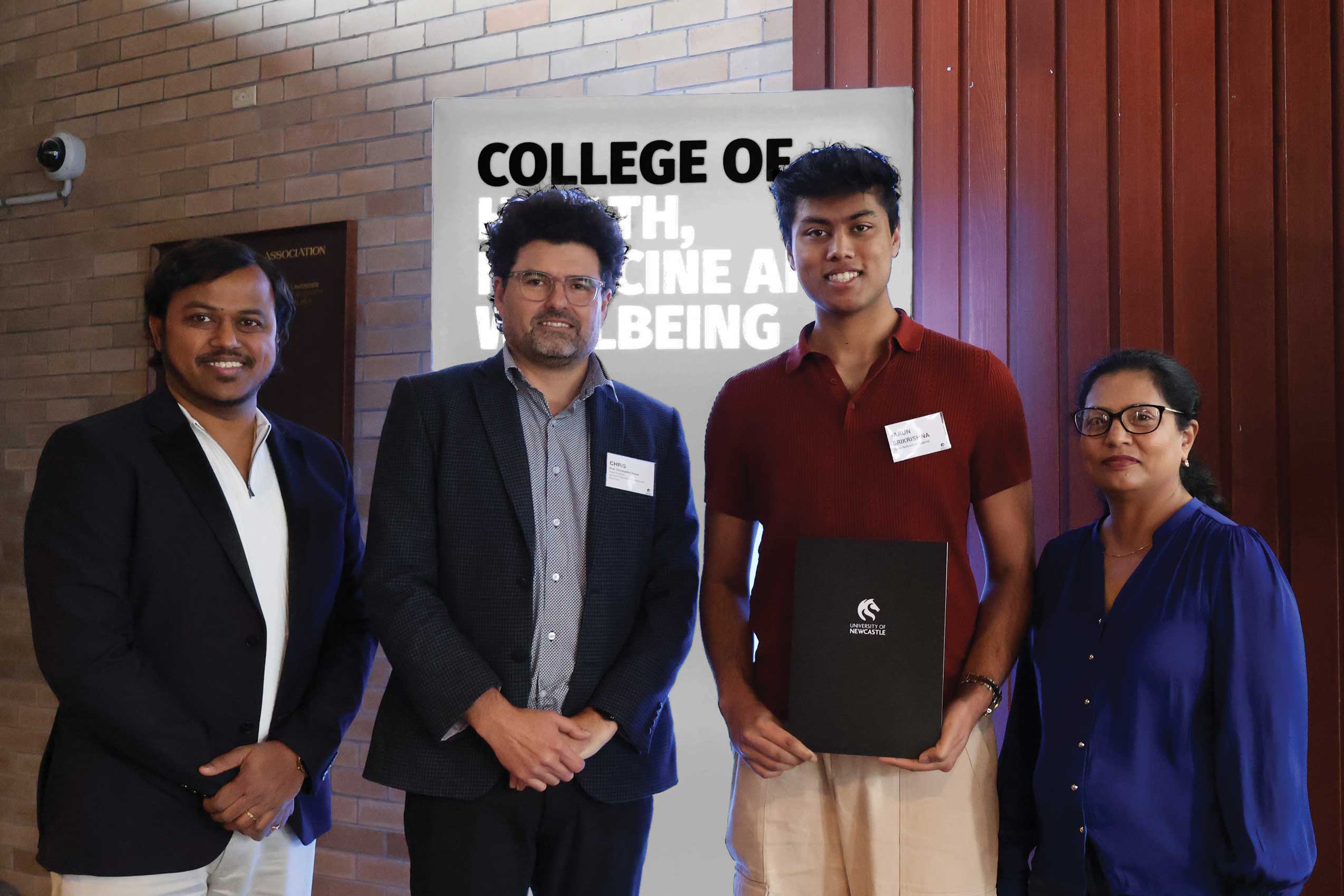 Four people stand together indoors in front of a sign reading “College of Health, Medicine and Wellbeing.” One person in the centre holds a black University of Newcastle folder, while the group poses for a photograph against a backdrop of brick and timber-panelled walls. All four are wearing name badges.
