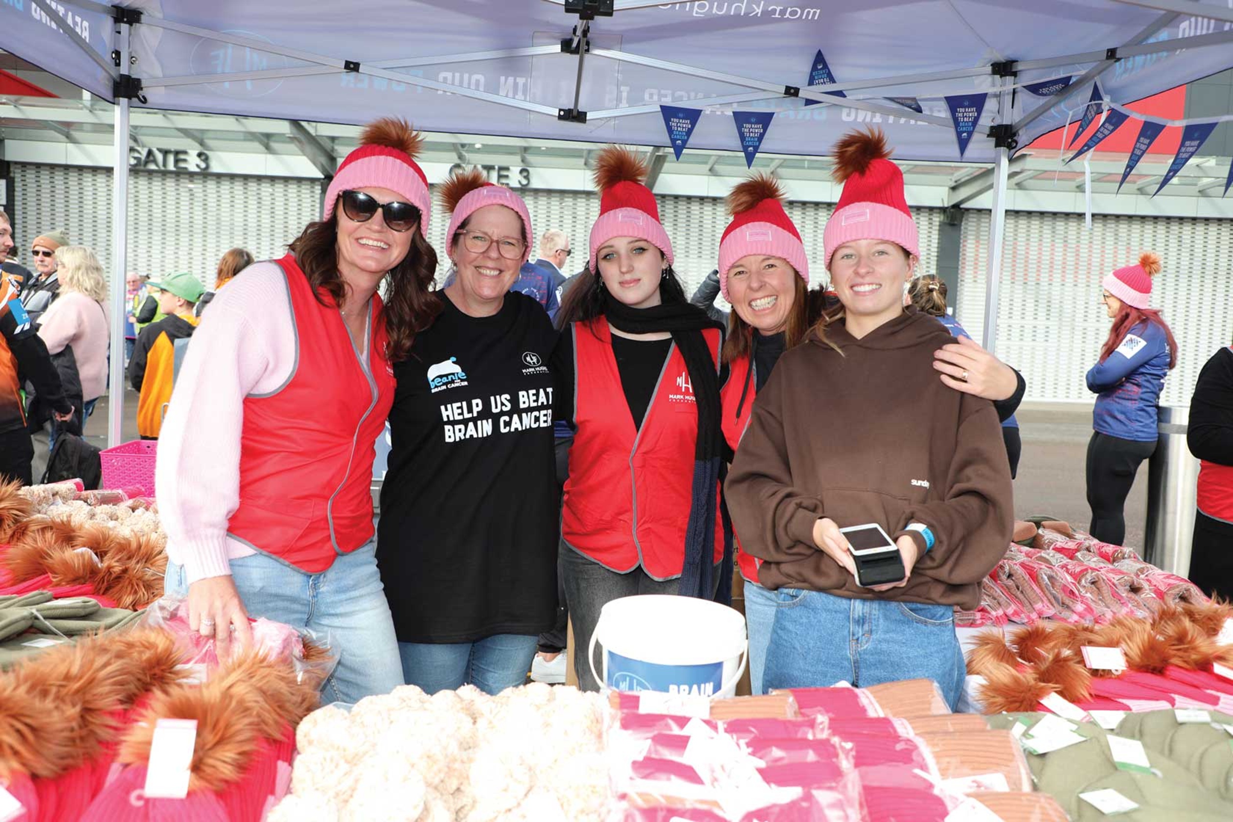 Five community members stand behind a fundraising stall under a marquee at a Newcastle Knights game. Several people wear bright pink beanies and red vests, with merchandise displayed on tables in the foreground. One person wears a shirt reading “Help Us Beat Brain Cancer,” and event attendees can be seen in the background.