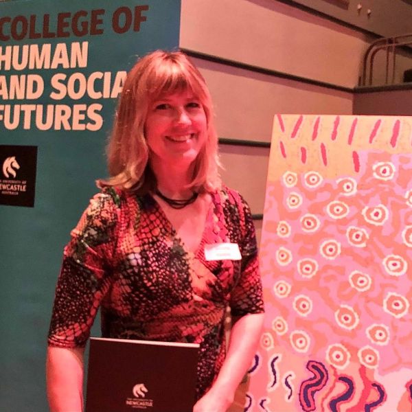 Woman smiling in front of Aboriginal artwork and University banner. 15-year milestone for Professor Victoria Haskins  