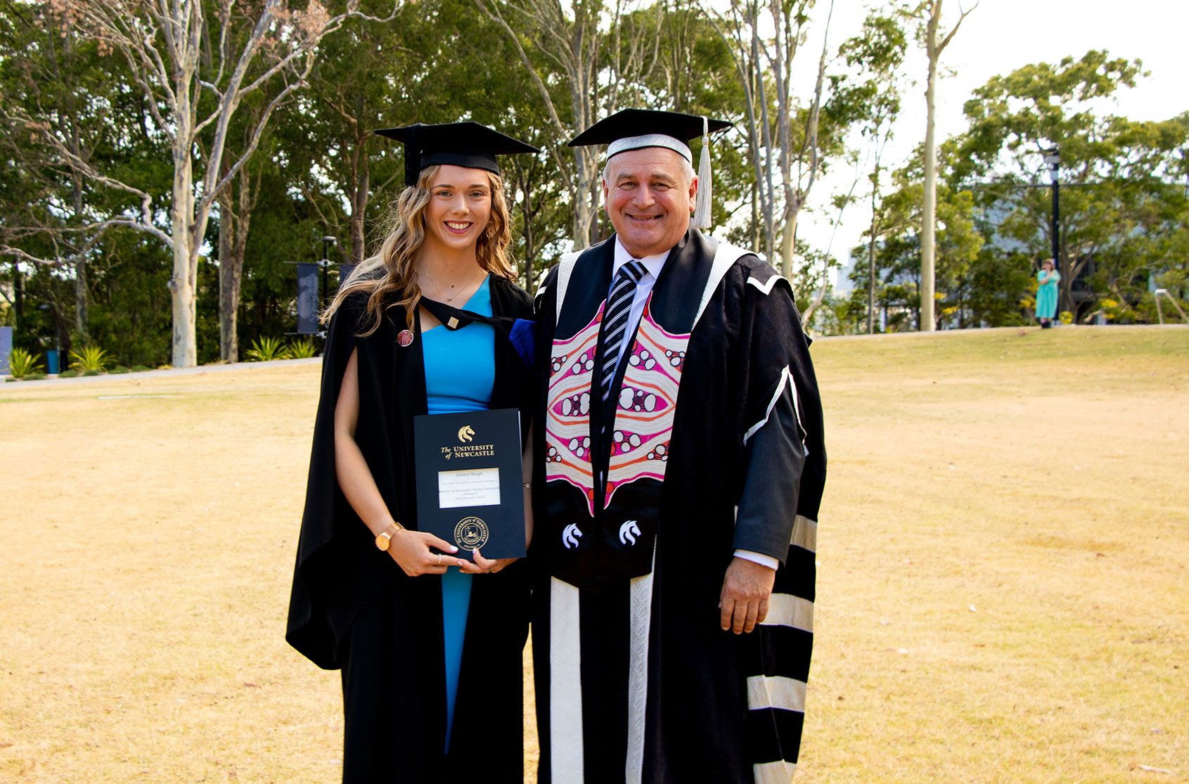 Jessica stands with her graduation certificate in her gown next to the Vice Chancellor in his official dress