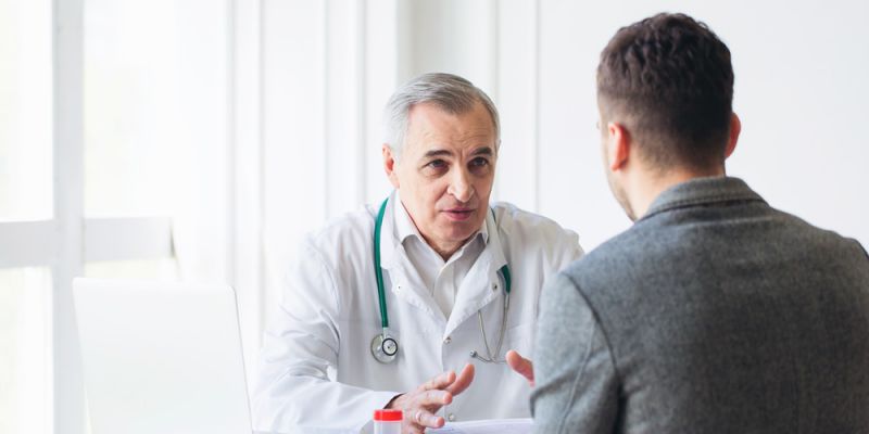 A male doctor sits at a table with a stethoscope around his neck. He is speaking to a patient.