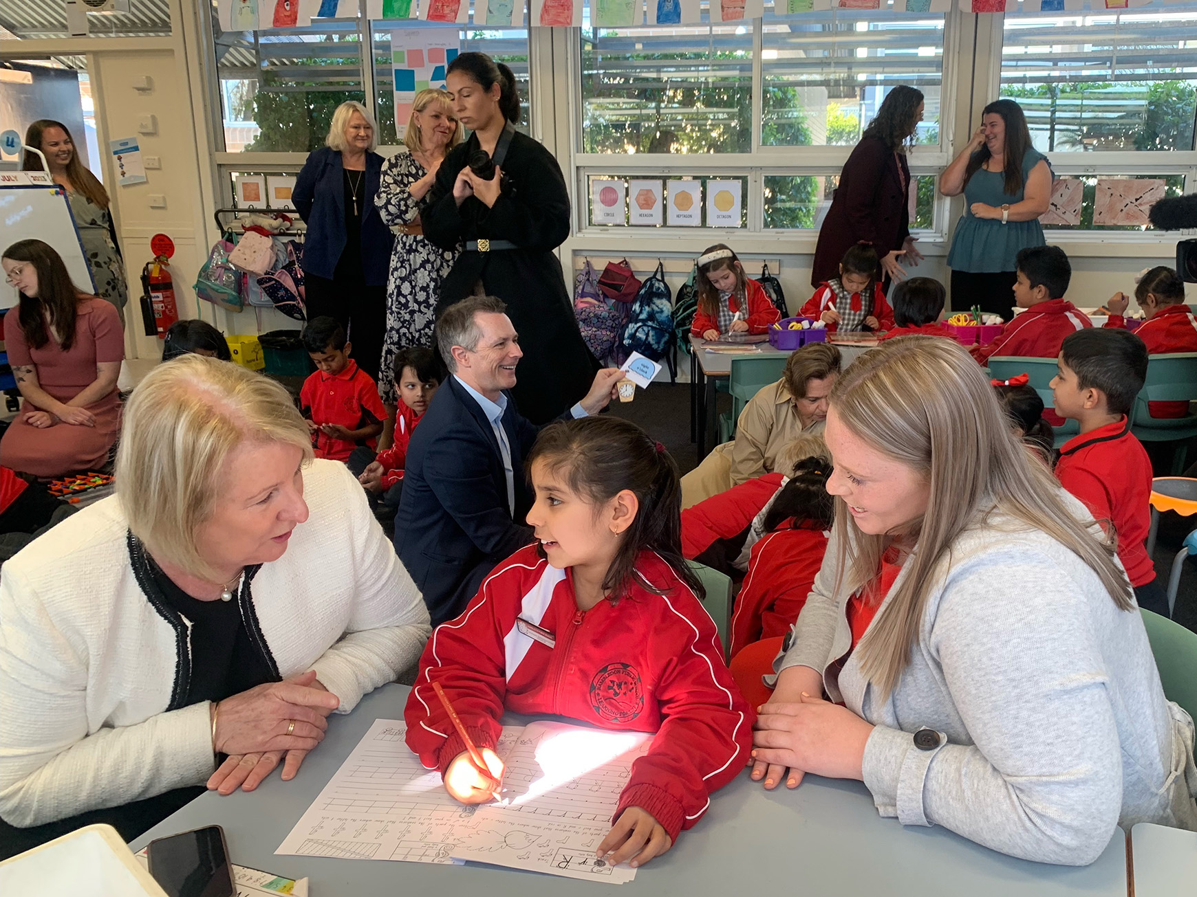 On the left a woman wearing a white jacket is talking to a primary school girl wearing a red uniform. On the right is a woman wearing a gray jacket. They are seated at a desk in a school classroom, there are other people and students working behind the. 