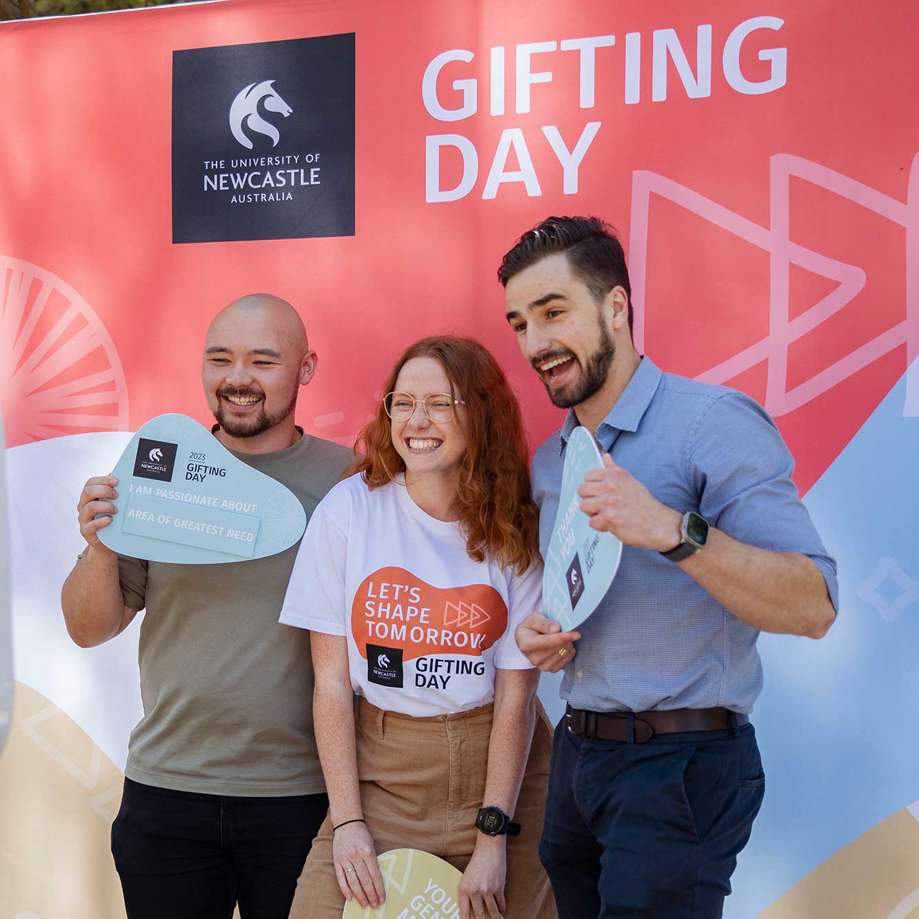 Image of three people in front of a photo booth screen, holding placards for Gifting Day