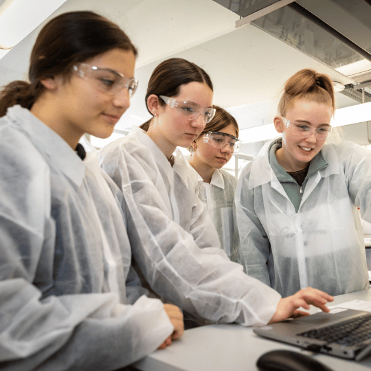 Four young women wearing lab coats and safety glasses are looking at a laptop in a laboratory^empty:{ds__assetid^as_asset:asset_name}