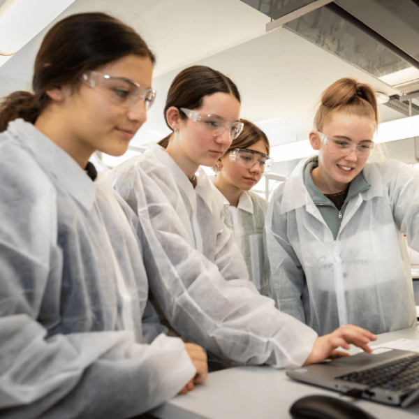 Four young women wearing lab coats and safety glasses are looking at a laptop in a laboratory