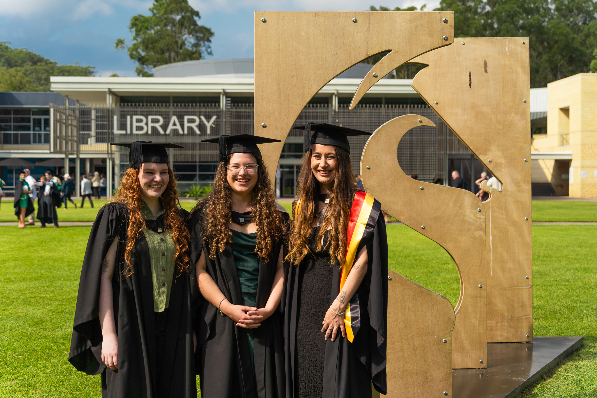 Three graduates smiling in front of Ourimbah campus library