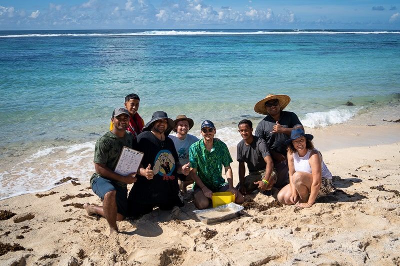 Team of Samoan and Australian scientists crouched on a beautiful Samoan beach smiling to camera with 'thumbs up'