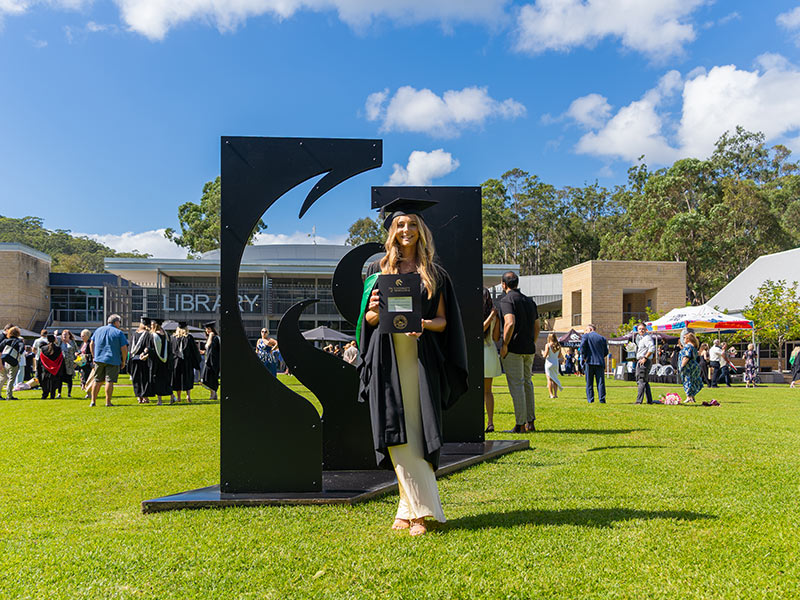 Piper Kennedy stands in front of the University logo statue proudly holding her degree and wearing cap and gown