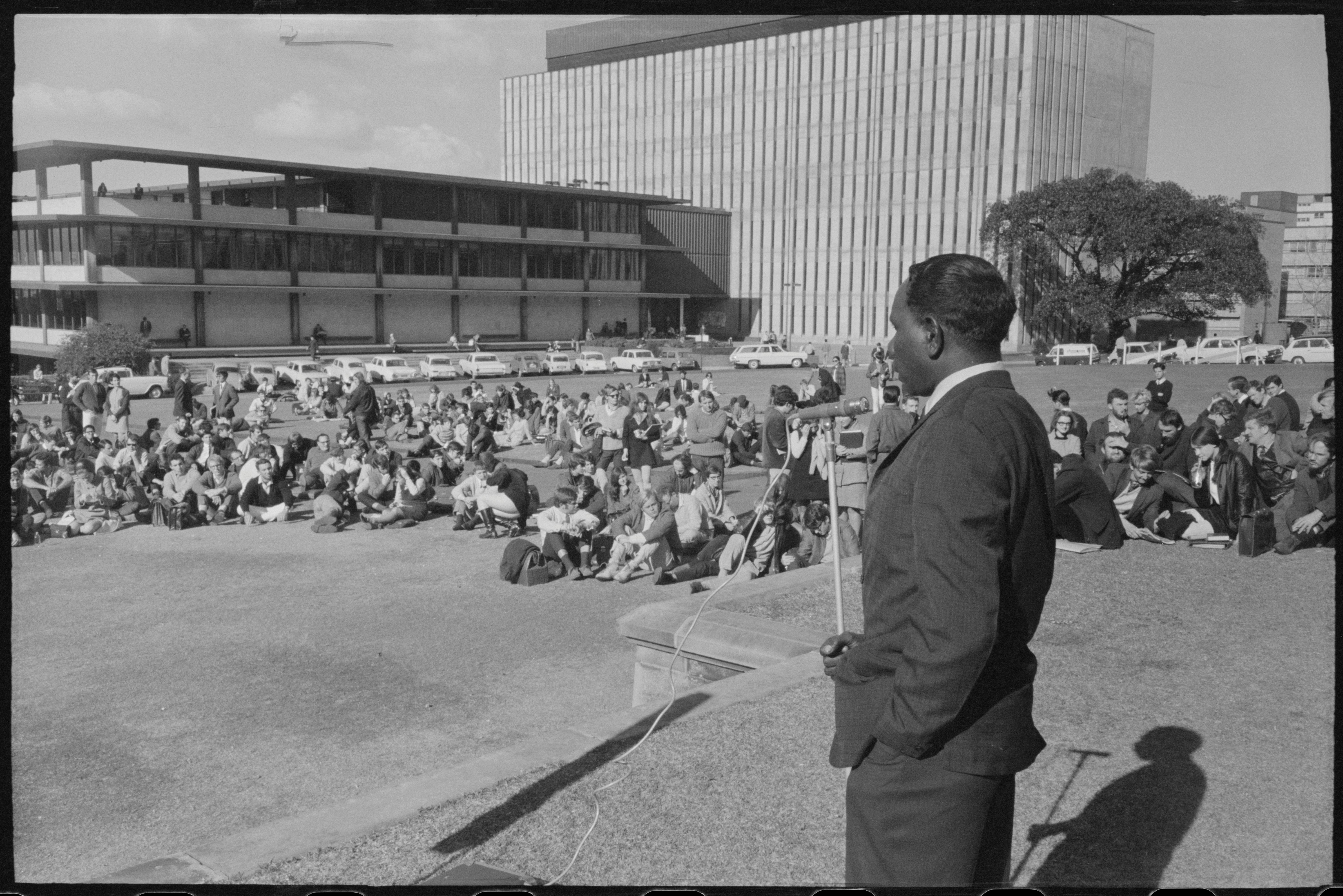 Dexter Daniels addressing university students (Supplied SLNSW) A B&W photograph of an Aboriginal man in a suit speaking into a microphone,addressing an audience of students sitting on the lawn