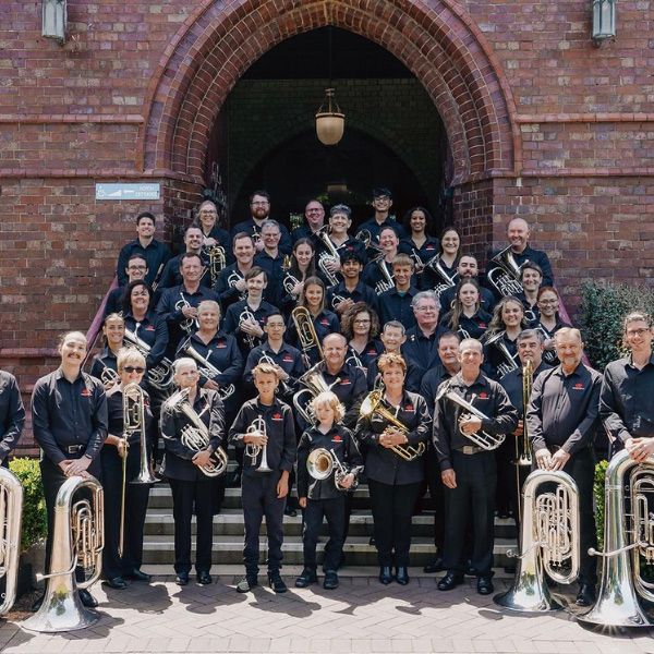 A large group in matching uniforms holds brass instruments while standing on the steps of a brick building with an arched entrance.