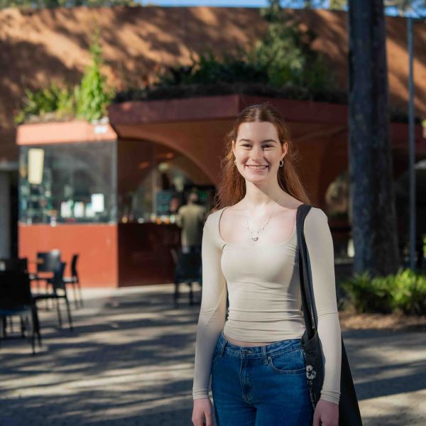 A young person stands outdoors in a sunlit area, smiling at the camera. They wear a light-colored long-sleeve top, blue jeans, and carry a black shoulder bag. Behind them is a red and brown cafe with large glass windows, surrounded by black chairs and tables. Trees cast shadows across the scene, and other people are visible in the background, sitting and walking around the University campus.