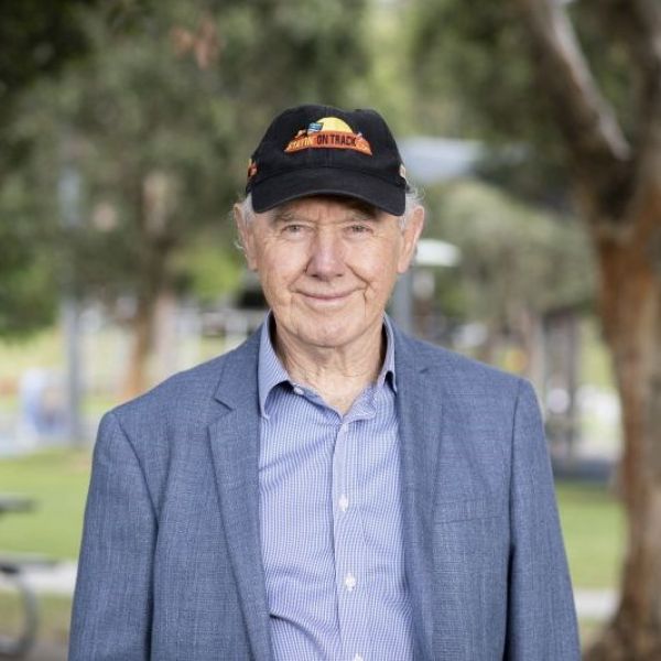 Man wearing baseball cap and suit is smiling for the camera with bushland in the background
