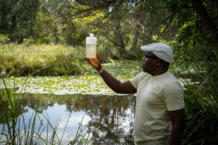 Simon inspecting a water sample Simon pictured holding up a water sample in a bottle. The backdrop is green and lush with a pond