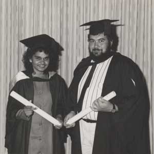 two students are shown smiling at the camera, in graduation gowns and caps, each holding a scroll