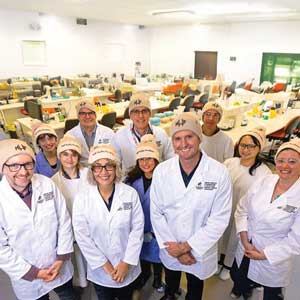 A group of people stand together with a laboratory behind them, all wearing lab coats and Mark Hughes Foundation beanies.