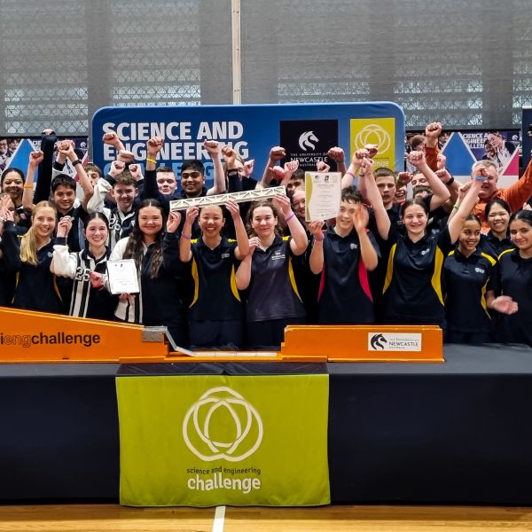 A large group of students wearing matching dark shirts with yellow accents stand behind a table, raising their arms in celebration. They are holding a certificate and a long bridge-like structure. Behind them is a banner that reads “Science and Engineering Challenge” with logos, and the table has a green cloth with the same event branding.. Science and Engineering Challenge celebrates National Finals.