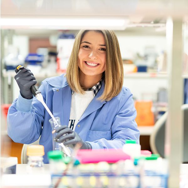 An image of a woman in a lab. She is smiling to camera and wearing a blue lab coat as she uses a pipette