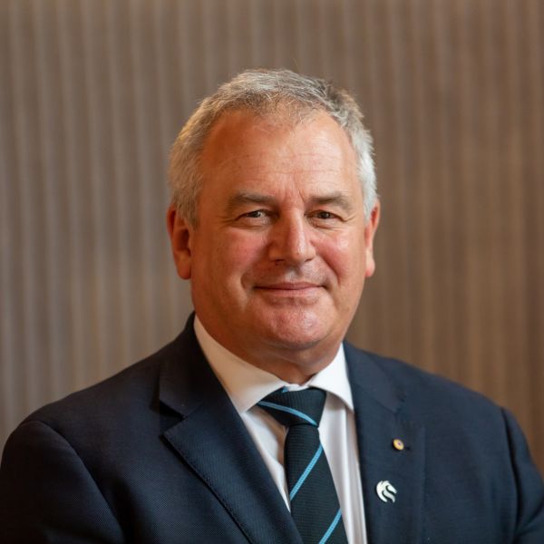 Vice Chancellor Alex Zelinsky is dressed in a dark suit jacket, white shirt and a striped tie, standing against a softly lit, vertically textured background. A small lapel pin of the University of Newcastle logo is visible on his jacket