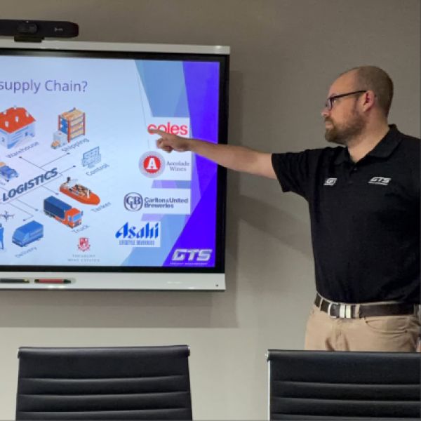 A man in a black shirt points at a presentation slide about logistics and supply chain. The slide features logos and illustrations of transportation trucks.