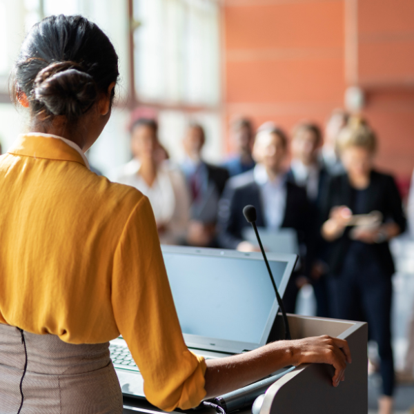 A person stands at a podium with a laptop, speaking to an audience gathered in a bright, modern conference space.