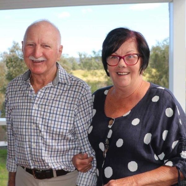 An older couple smiling and looking at the camera, standing on a balcony overlooking a lake. . Why I donate - Cheryl and Barrie Graves.