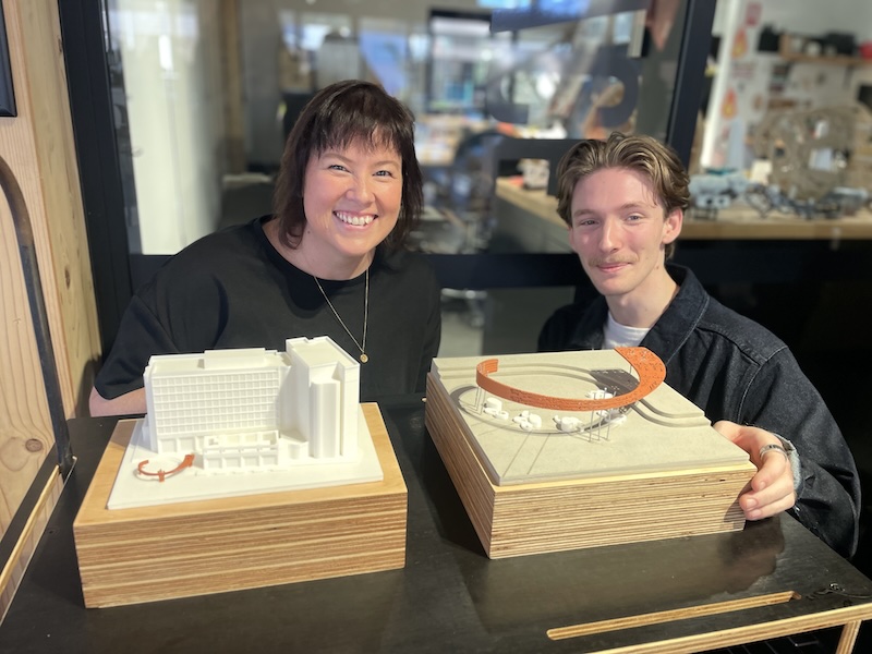 Thirs year Architecture stduent Sarah Hurley and Masters of Architecture student Sam Orme with a model of the design of Turntable Plaza. A young woman and a young man smile and pose behind a model of a building.