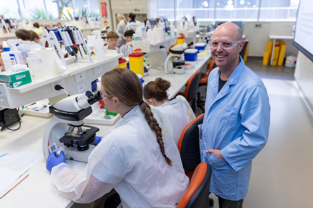 High school student wearing a lab coat and protective goggles uses a microscope, supervised by a male Academic staff member who is smiling at the camera