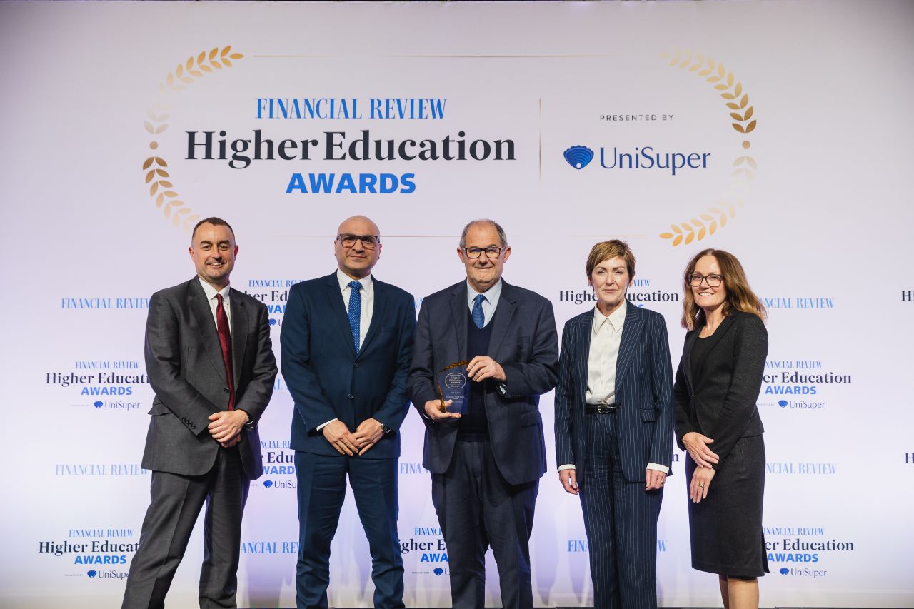 Five people, three men and two women stand in a line on a stage looking at the camera. The AFR Higher Education Awards banner is in the background. The man in the middle is holding an award.