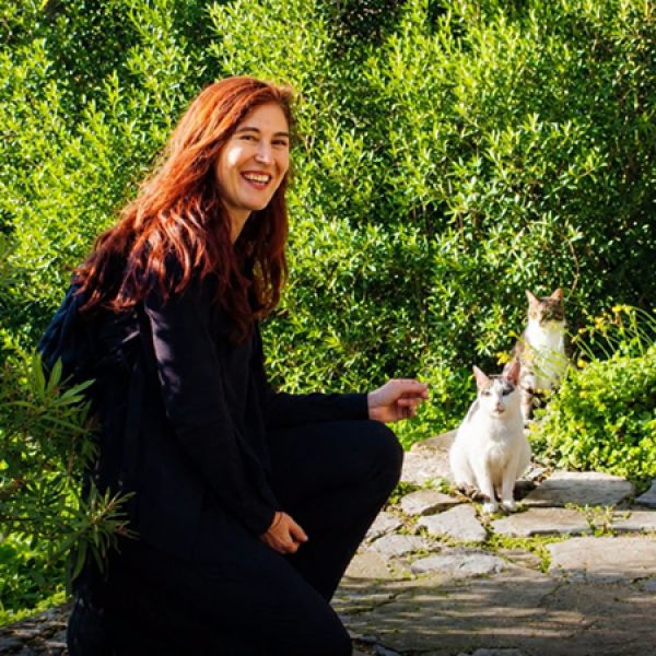 Person crouching on a stone path in a lush green garden, reaching toward two cats sitting nearby among the foliage.