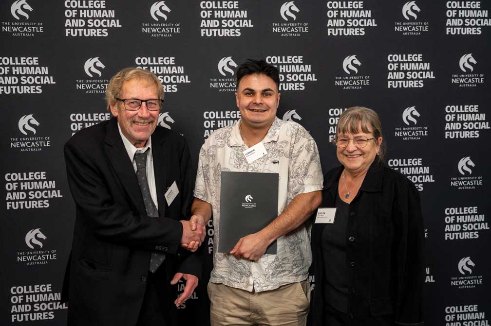 Three people stand in front of a University of Newcastle College of Human and Social Futures media wall. One person in a suit shakes hands with a student holding a University of Newcastle certificate folder, while a third person stands alongside. The group poses for a photo at a recognition or award presentation event.