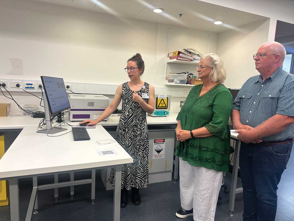 Three people stand in a laboratory, with Dr Heather Murray presenting research findings on a computer monitor. Laboratory equipment and workstations surround the group, while Ngaire and Bruce Callaghan observe the demonstration.