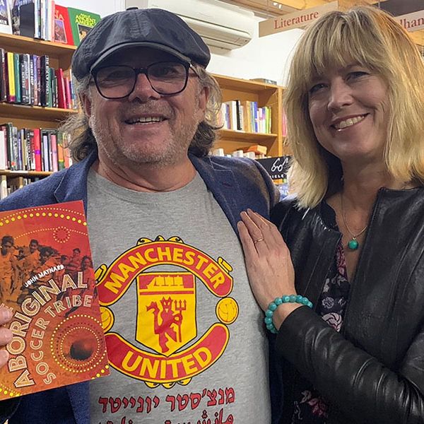 John Maynard and partner, Victoria Haskins smiling in bookshop holding John's book. Kicking Goals for Aboriginal Soccer 