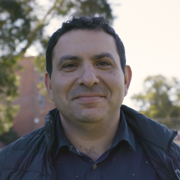A close-up picture of Sam, a participant in the Mid-Career Transition to Teaching Program. He is wearing a blue collared shirt and a black jacket with trees in the background