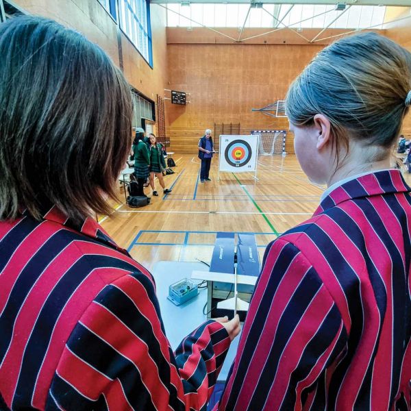 Two students in blazers with their backs to the camera, watching a glider head towards a target in a sports hall