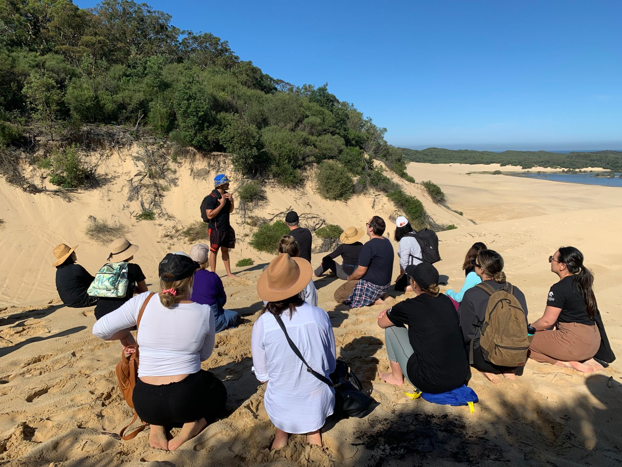 A group of people sit in a circle on sand dunes listening to the speaker who is standing^empty:{ds__assetid^as_asset:asset_name}