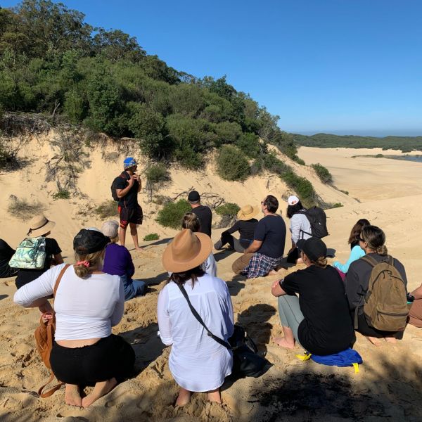 A group of people sit in a circle on sand dunes listening to the speaker who is standing