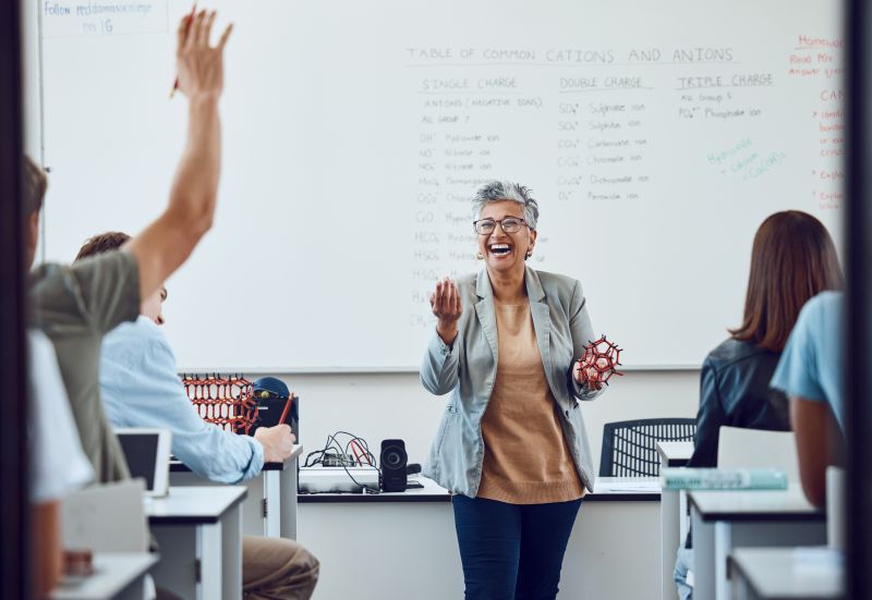 female teacher in grey blazer stands at front of a classroom gesturing at a student with their hand up