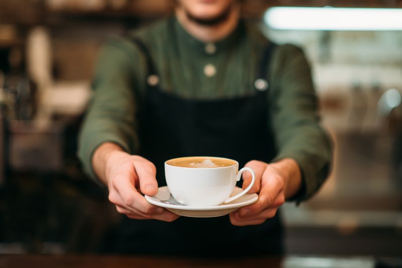 Male barista in green shirt and black apron, holding out a cup of coffee sitting on a saucer