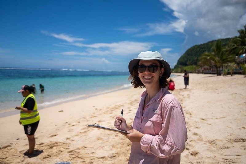 Female student, Courtney, smiling at the camera holding a clipboard taking notes on a beautiful beach