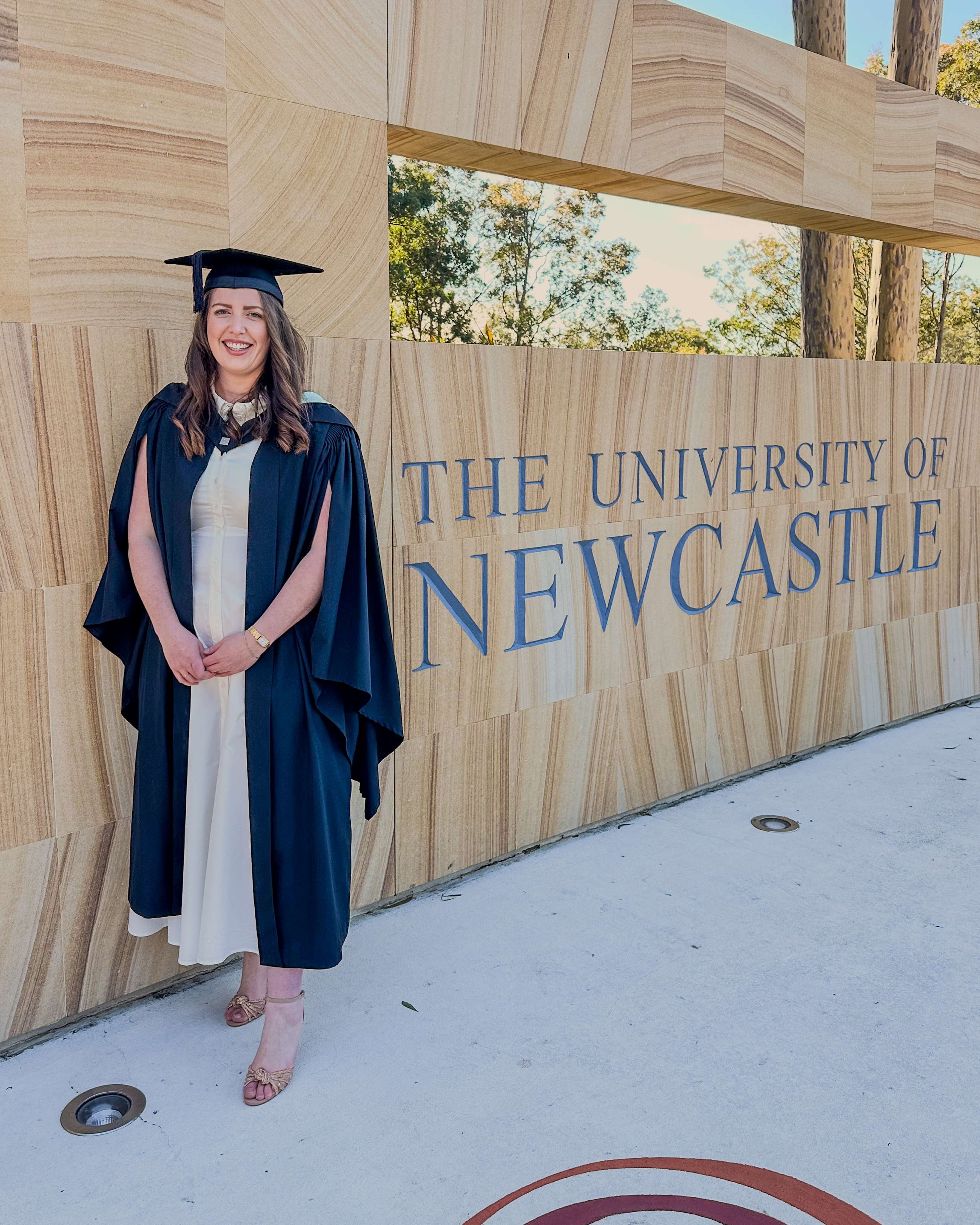 Rebecca Cramp in her graduation robes standing in front of a University of Newcastle sign